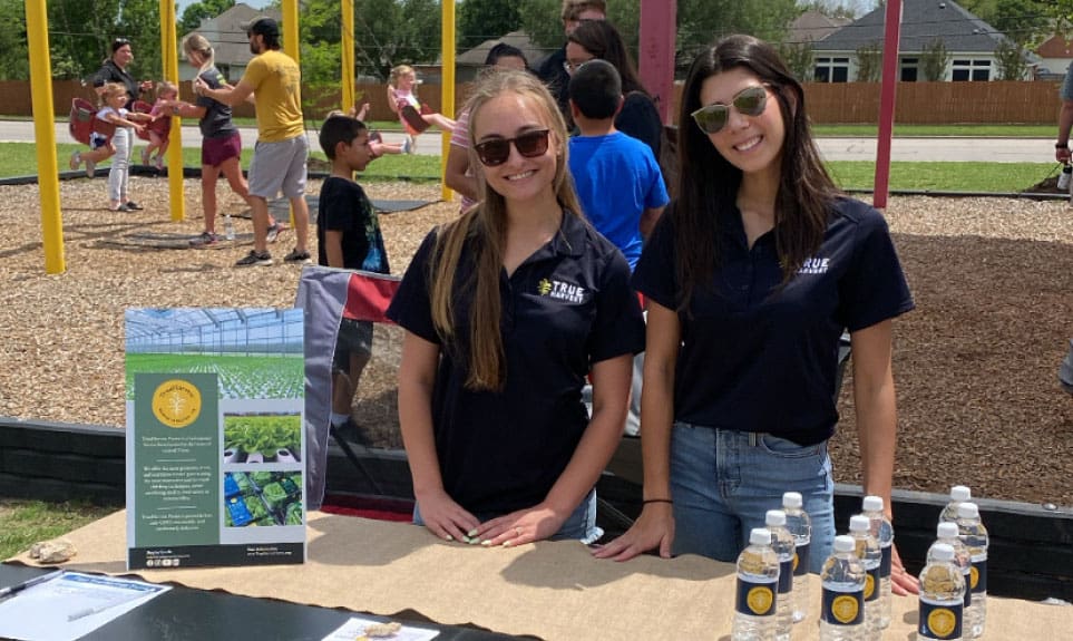 Two women standing together in TrueHarvest apparel smiling outside at a promotional table.